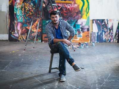 Portrait of young artist painter sitting in front on his big modern paintings in art studio.