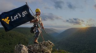 WVU student holding a West Virginia University flag that reads "Let's Go." standing on top of a mountain.