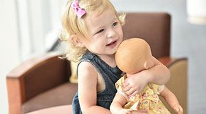 Blonde, female toddler holding a doll in front of a chair.