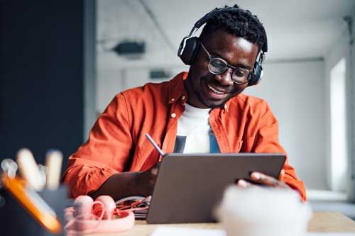 Happy African-American student sitting at desk with his unrecognizable friend while listening online lesson on his digital tablet and writing homework in a notebook.