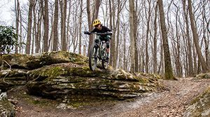 Young girl jumping a bike on a trail over a large rock.